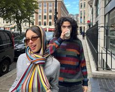 two people walking down the street, one holding a coffee cup in her hand and the other wearing a scarf around her neck