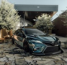 a black car parked in front of a building on a cobblestone driveway with trees and bushes