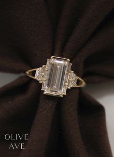 an emerald cut diamond ring sitting on top of a brown cloth with the words olive ave written