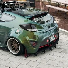 the rear end of a green car parked in front of a park bench on a brick sidewalk