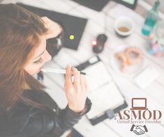 a woman sitting at a desk with a pen in her mouth and an electronic device next to her