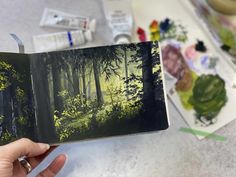 a person holding up an open book in front of some paintbrushes and other art supplies