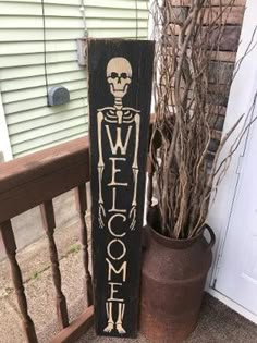 a welcome sign sitting next to a potted plant on the front porch with a skeleton painted on it