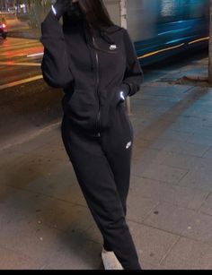 a woman is standing on the sidewalk talking on her cell phone while wearing a black tracksuit