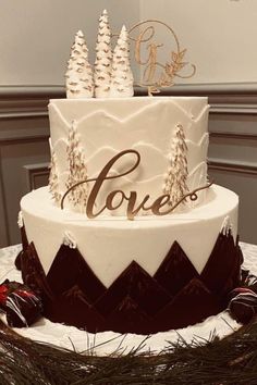 a white and brown wedding cake with pine trees on the top is decorated with gold lettering