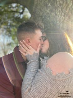 a man and woman kissing in front of a large tree with the sun shining down on them