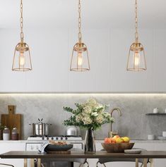 three lights hanging over a kitchen island with fruit on the counter and flowers in vase