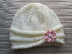 a white knitted hat with a pink flower on the front and side, sitting on top of a wooden table