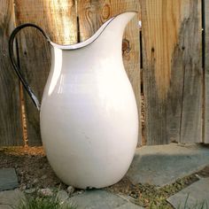 a white pitcher sitting on the ground next to a wooden fence