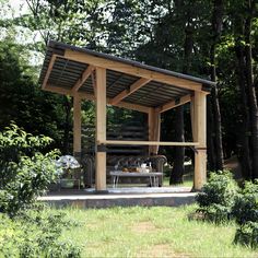 a wooden structure in the middle of a forest with benches and tables under it, surrounded by trees