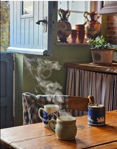 a wooden table topped with two mugs filled with steaming liquid next to a window