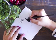 two hands writing on paper next to a potted plant and bowl of cherries