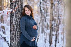 a pregnant woman standing next to a tree in the snow