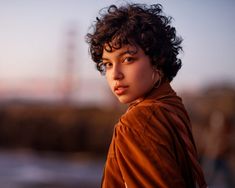 a close up of a person wearing a brown jacket and large hoop earrings, looking at the camera