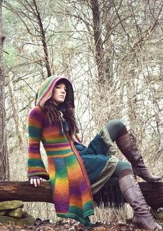 a woman is sitting on a log in the woods wearing a colorful sweater and boots