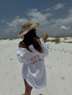 a woman in a white dress and straw hat on the beach drinking from a cup