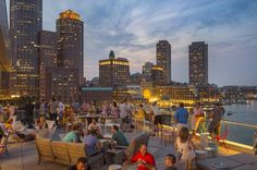 many people are sitting at tables on the roof of a building with city lights in the background