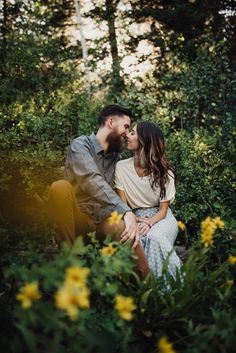 a man and woman sitting on the ground in front of some trees with yellow flowers