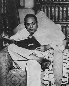 a black and white photo of a man sitting on a chair reading a book in front of bookshelves