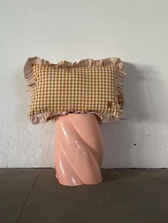 a pink vase sitting on top of a floor next to a white wall with a brown checkered pillow