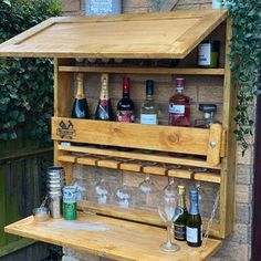 a wooden shelf with bottles and glasses on it in front of a brick wall next to a bush