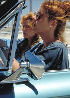 two women sitting in the driver's seat of a blue car, one with her hand on the steering wheel