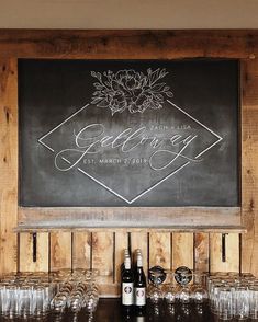 there is a chalkboard sign on the wall above some wine glasses and bottles in front of it