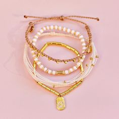 three bracelets with gold charms and beads on a pink background, one has a charm