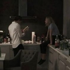 a man and woman standing in a kitchen next to a counter with bottles on it