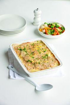 a casserole dish on a table next to a bowl of vegetables and a fork