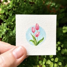 a hand holding up a small card with pink flowers on it in front of some green plants