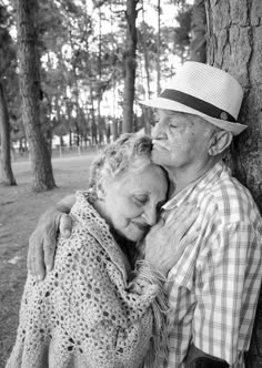 an old man and woman embracing each other in front of a tree with their arms around one another