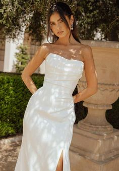 a woman standing in front of a stone wall wearing a white dress with high slit