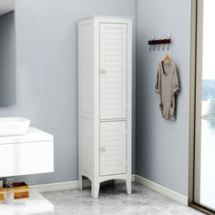 a white cabinet in a blue bathroom next to a mirror and door with clothes hanging on it