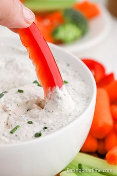 a person dipping dip into a white bowl with carrots and celery in the background