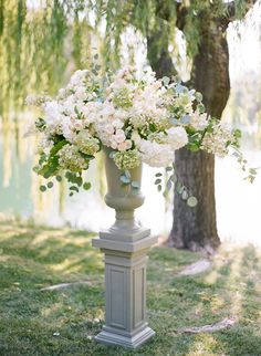 a tall vase filled with white flowers sitting on top of a grass covered field next to a tree