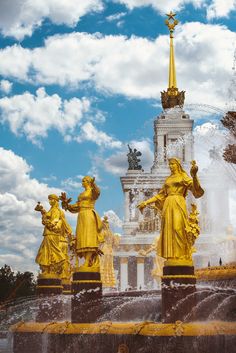 three golden statues in front of a fountain with blue sky and white clouds behind them