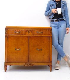 a woman standing next to a wooden cabinet and holding a coffee cup in her hand