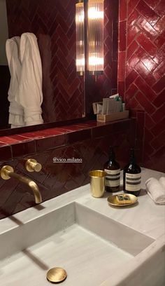 a white sink sitting under a bathroom mirror next to a red tiled wall and gold faucet