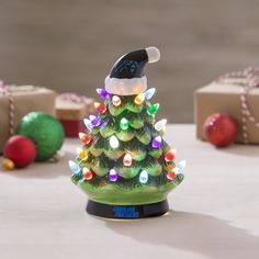 a small christmas tree with lights and a santa hat on it's head is sitting in front of presents