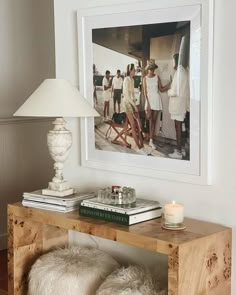 a wooden table topped with books next to a white lamp and framed photograph on the wall