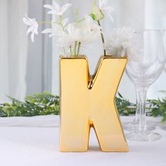 a gold letter shaped vase with flowers in it next to two wine glasses on a table