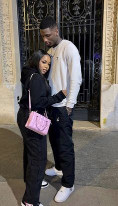 a man standing next to a woman in front of a gate with a pink purse