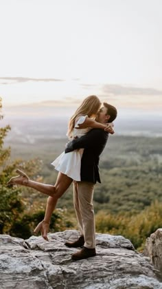 a man and woman are kissing on top of a rock in the mountains at sunset