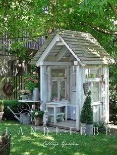 a small white shed sitting in the middle of a lush green yard with potted plants