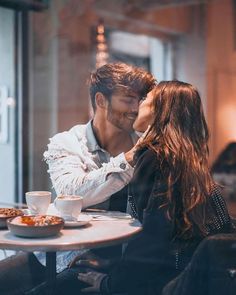 a man and woman sitting at a table with coffee in front of them, kissing