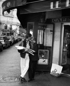 black and white photograph of two people kissing in front of a cafe with cars parked on the street