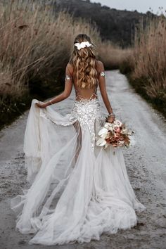 a woman in a wedding dress walking down a dirt road with her back to the camera