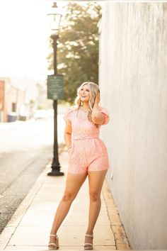 a woman is posing on the sidewalk with her hand in her hair and wearing a pink romper