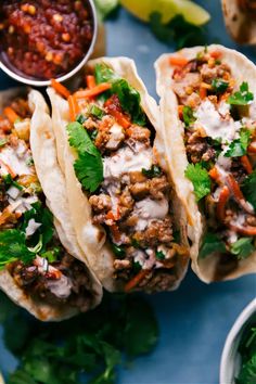 three tacos with meat, vegetables and sauces on a blue plate next to lime wedges